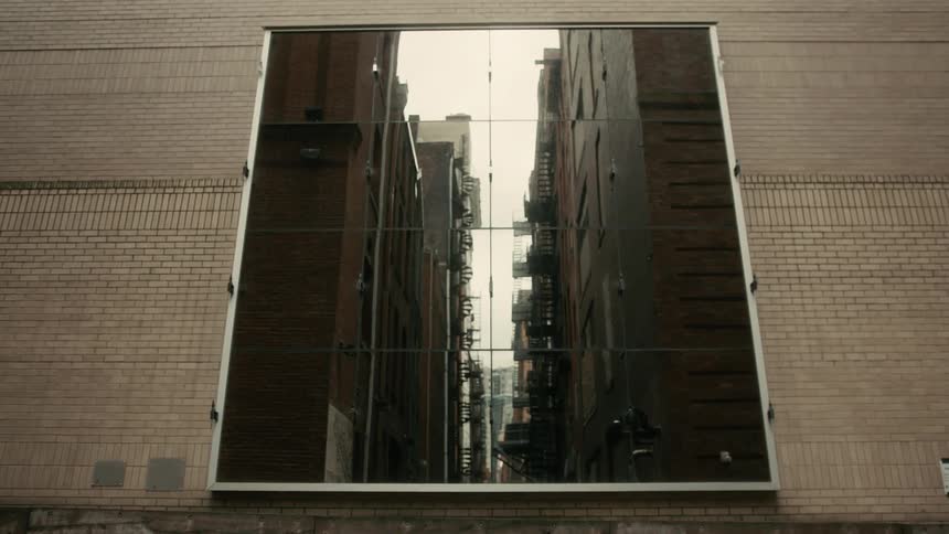 Large digital rectangular mirror mounted on a brick wall reflecting the view of a narrow alley between buildings