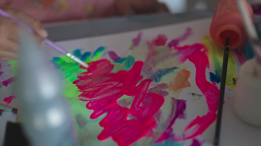 A girls hand holds a paintbrush applying bright pink paint to a paper with colorful streaks and splatters