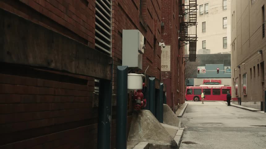 People mill about near a city alley and a street with bus and vehicle traffic