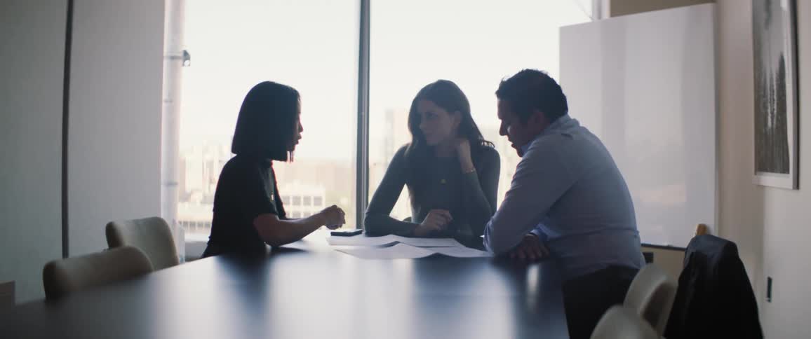 A meeting of three people around a black office table and behind is a large glass window
