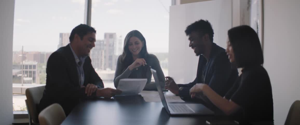 Two men and two women are gathered at a table looking at papers and a notebook and a man is smiling