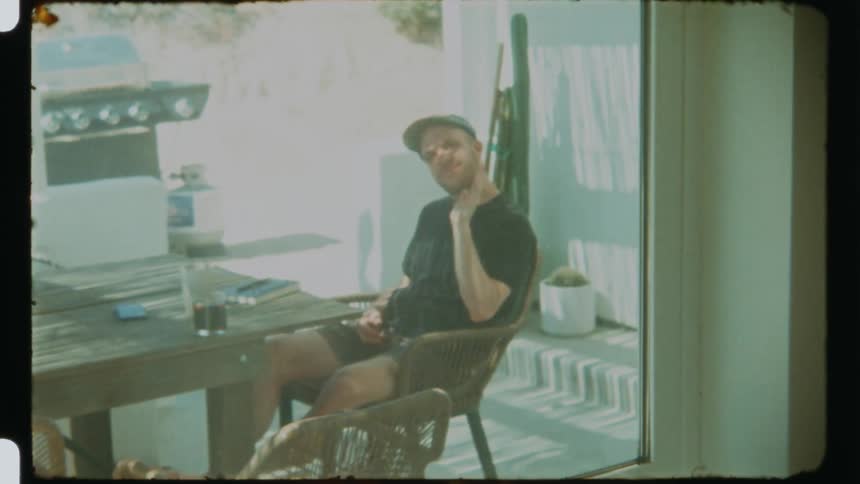 A man sitting on a balcony is waving and there is a glass window in front of him