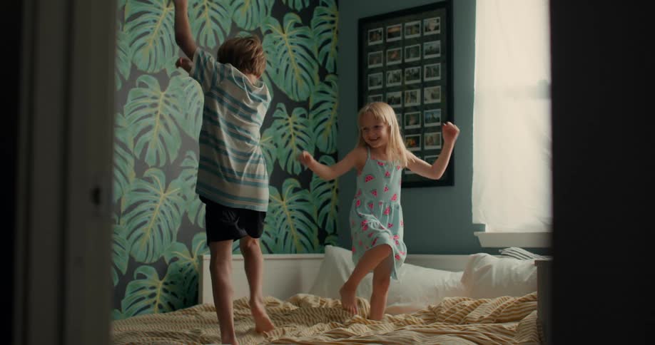 Brother and sister playfully jumping and dancing in a bed on a colorful bedroom