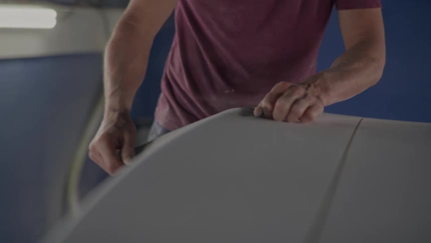A closeup of a persons hands sanding the side of a surfboard in a workshop