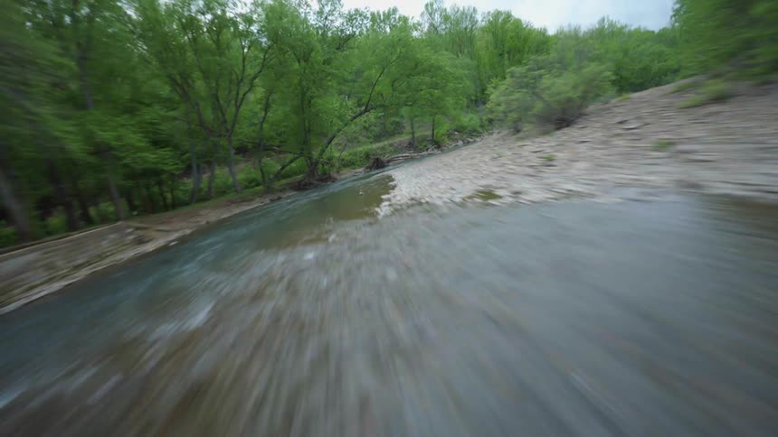 A stream with a rocky bank cuts through a forest under fluffy white clouds
