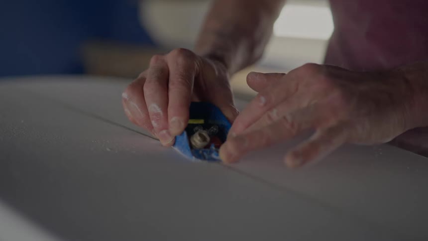 Closeup of two hands working on a surfboard with a tool used to shape and repair the board