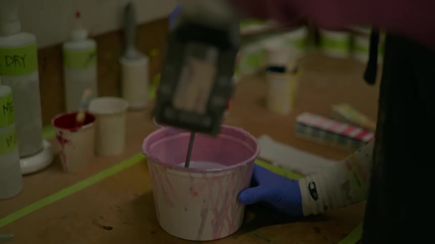 Hand with blue gloves holding a pink container with a viscous substance and the other hand using a tool in the liquid