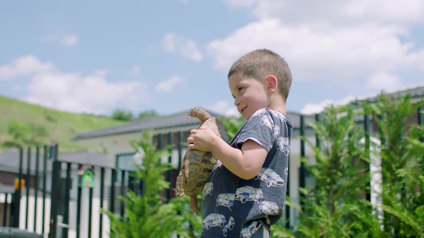 Boy smiles while carrying a tortoise through the backyard on a cloudy day