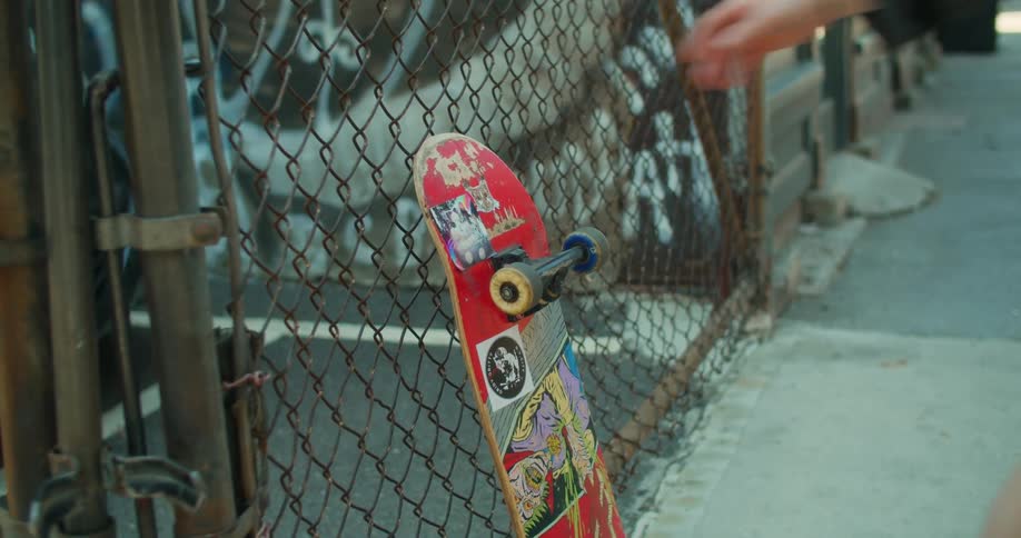 A white female grabs a skateboard thats leaning against a fence in a city