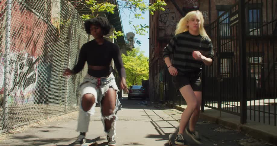 A black girl and a white girl skateboard in an alley