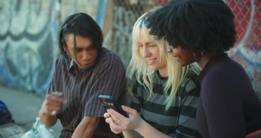 Two women and a man are sitting and looking at a cell phone while laughing and talking