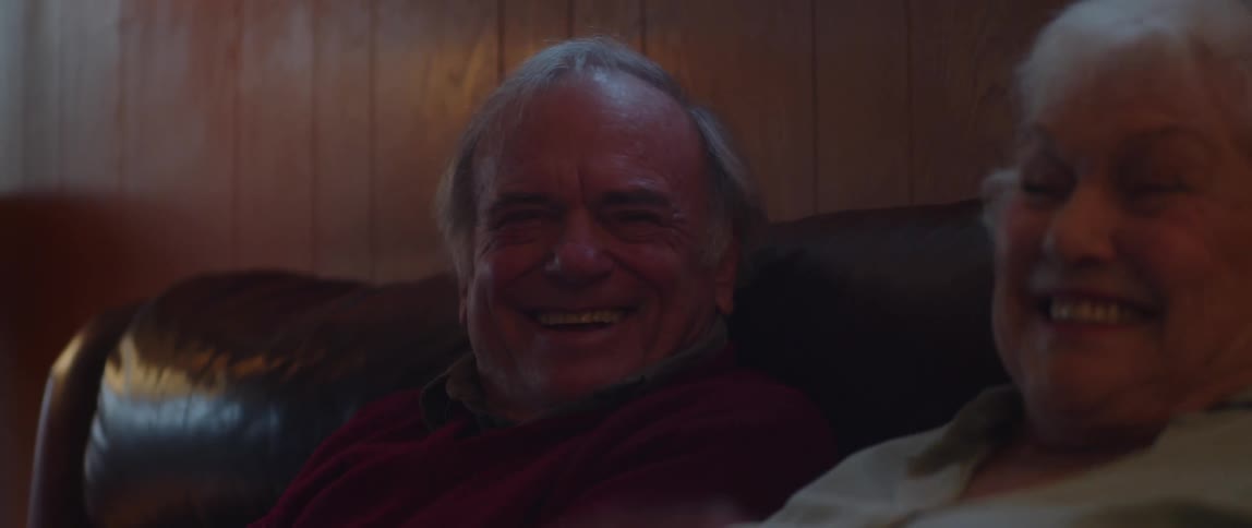 An elderly couple laugh while watching something while sitting on the couch