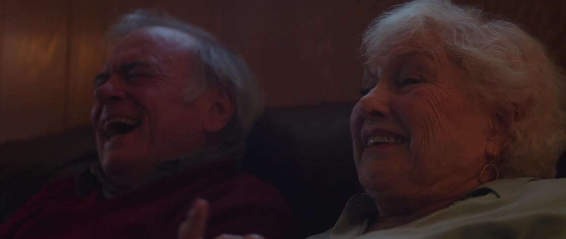 An elderly couple sitting on the couch at home laugh while watching television together