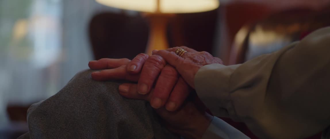 An elderly couple hold hands while sitting in a living room