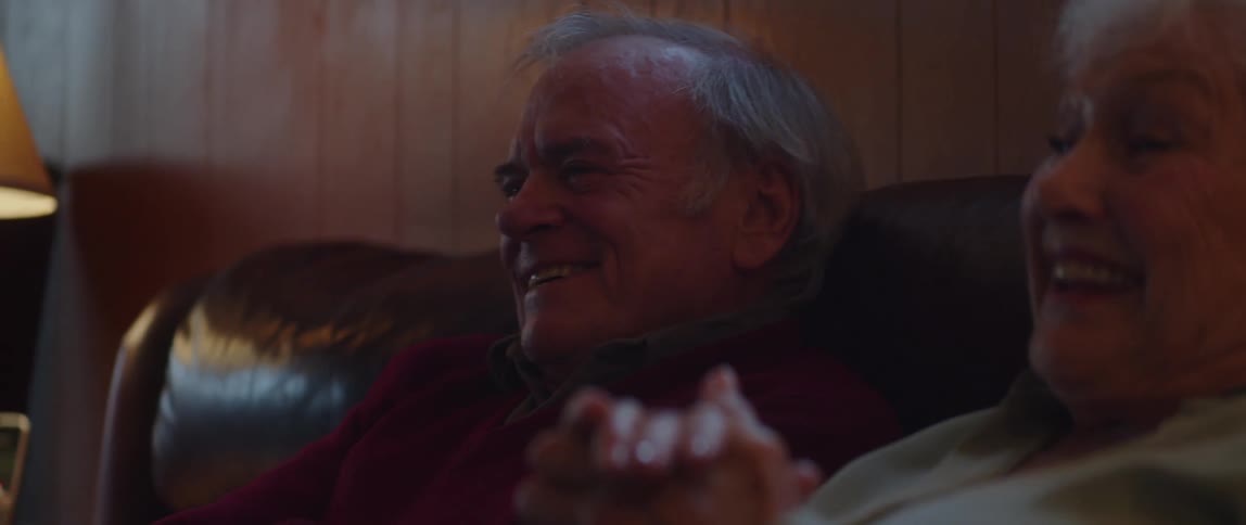 Elderly couple sits on a couch laughing while enjoying a broadcast