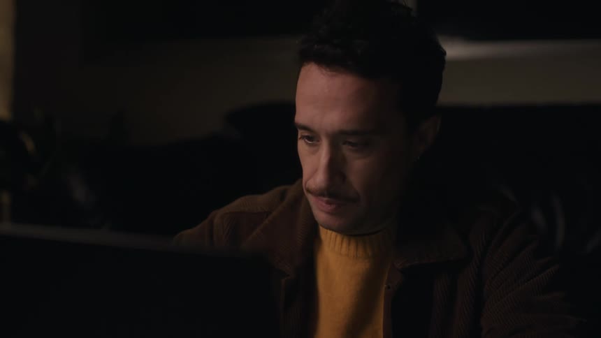 Man sits in a dimly lit room and smiles while using a laptop