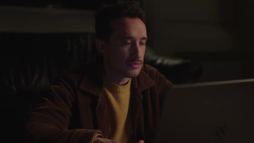 A man wearing a brown jacket and a yellow sweater sits in a dimly lit room focused on a laptop