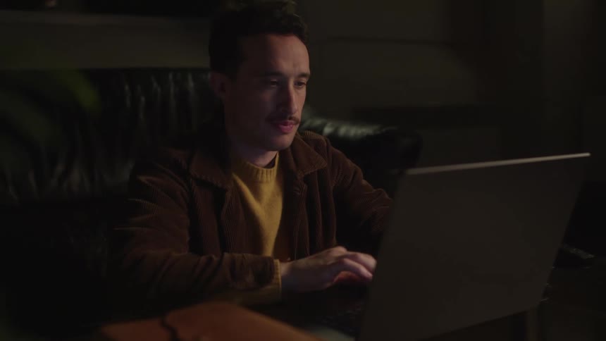Man stretching while sitting and working on a laptop in a room with low lighting