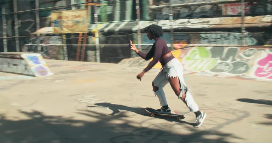 Woman rides a skateboard while using a smartphone to video her friend on a second skateboard at a skate park
