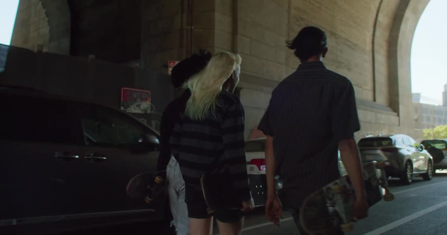 Three skater friends walk down a covered city street while holding skateboards