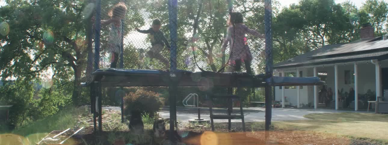 Three kids jump on a trampoline in a backyard as two dogs bark and run around underneath the trampoline