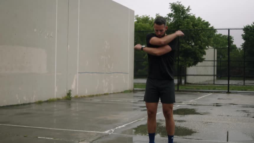 A young bearded man performing warm up exercises on a wet court under an overcast sky