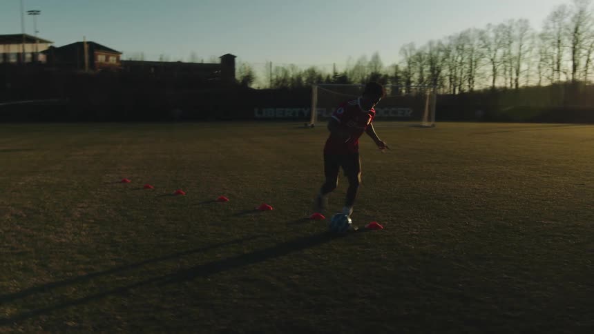 A person dribbling a soccer ball through orange cones on a grassy field