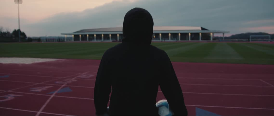 An individual walks onto a field holding a soccer ball and some shoes