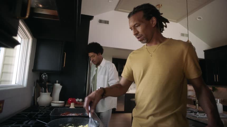In a kitchen a man whisks eggs while another chops tomatoes