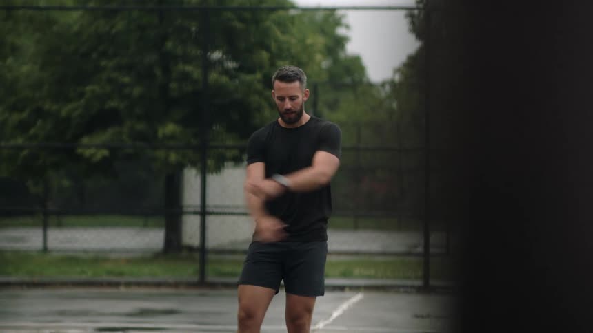 A young bearded man performing warm up exercises on a wet court under an overcast sky