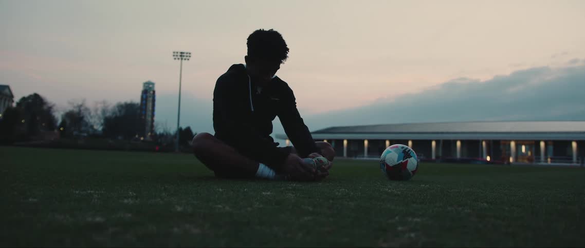 Boy sitting on the grass next to a soccer ball stretching his legs