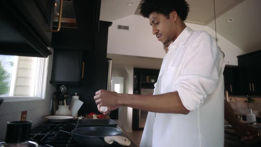 Two men in the kitchen with one of them pouring and cracking an egg into the frying pan