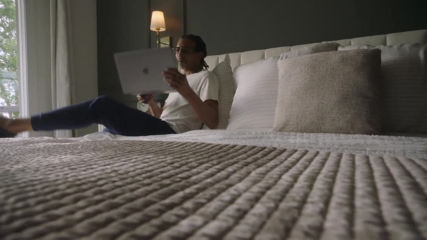 A man sits on his bed and uses a laptop