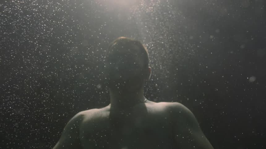 A man is under the dark water and he is blowing bubbles out of his mouth