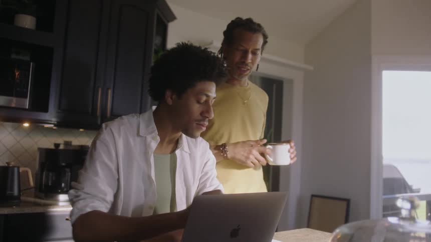 Two men in the kitchen with one of them sitting working on a laptop