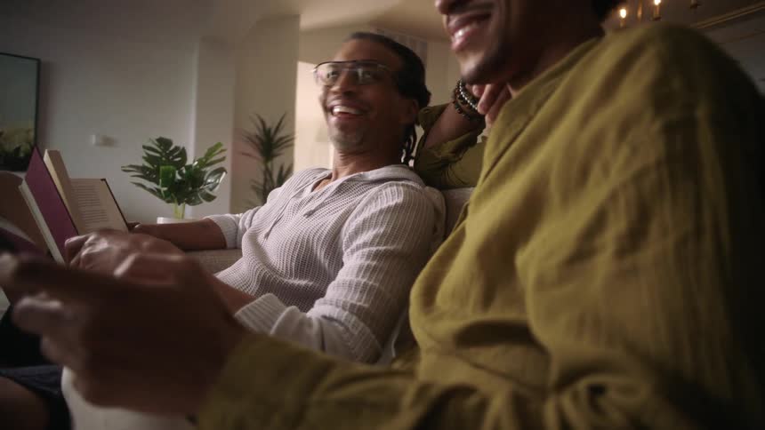 Two smiling men sitting in the living room with one of them reading a book and the other using a tablet