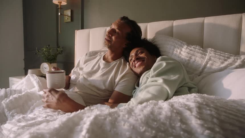 Two men are lying in bed hugging each other laughing and talking and one of them is holding a mug of hot liquid