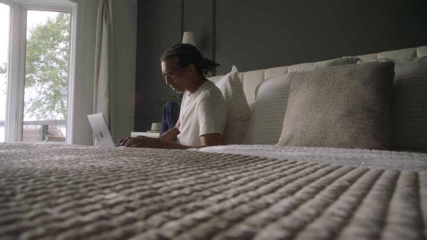 A man sitting on a bed with a laptop in a room with natural light coming through a window