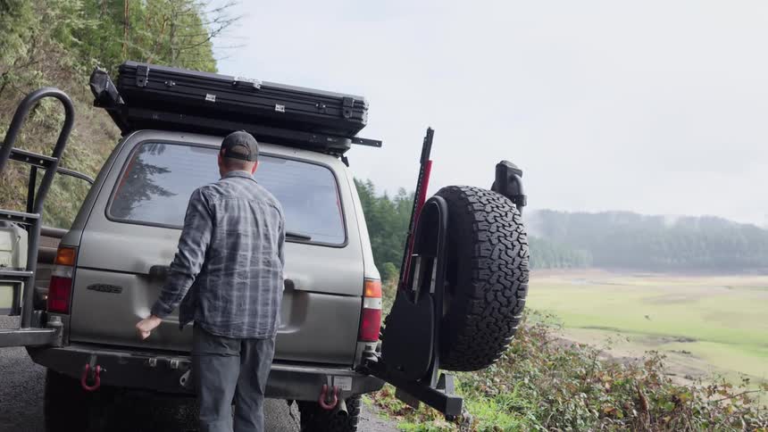 A man opens the trunk of a silver offroad vehicle parked on a dirt road in the countryside