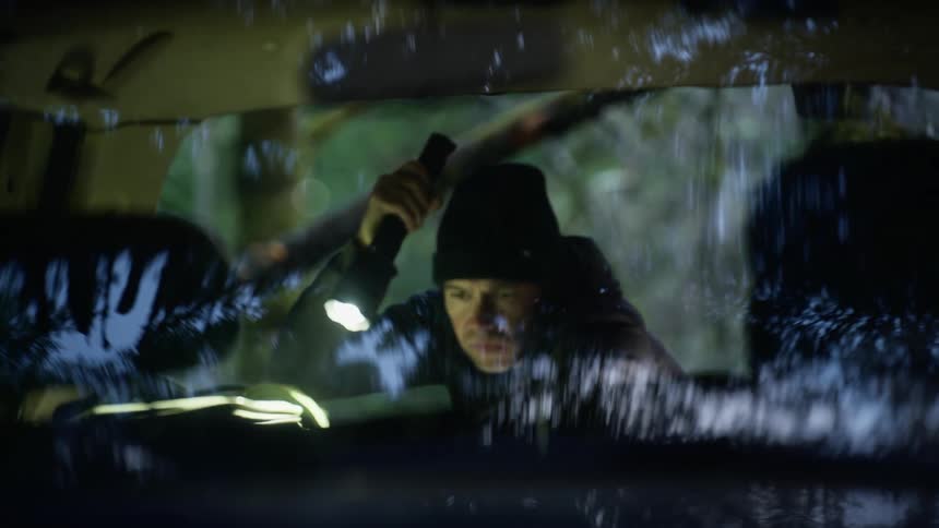 A man wearing a hoodie searches the interior of a vehicle with a flashlight