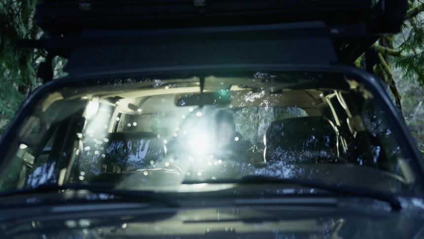A man uses a flashlight to search the trunk of a vehicle parked in the forest