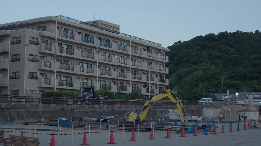 A multistory building stands behind a construction site with an excavator and orange traffic cones