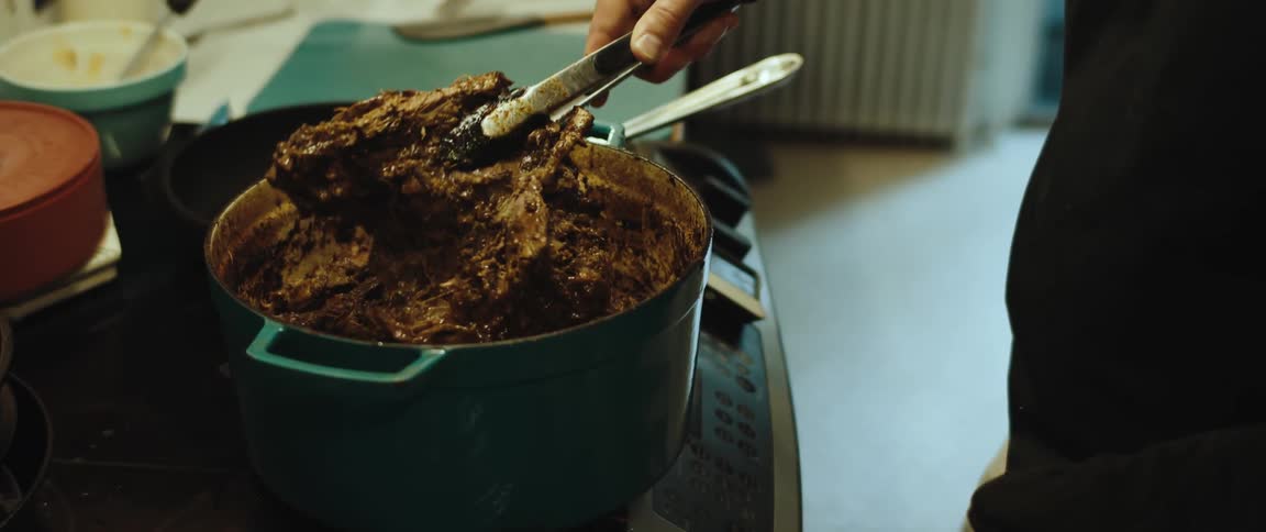 A man using tongs to pick up a dark stew cooking inside a large cooking pot on a stove