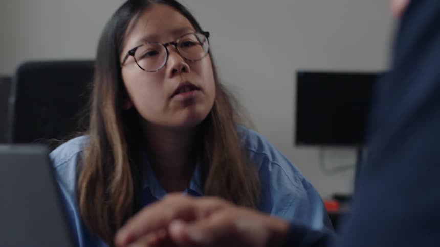 A young woman talks to another individual at a business meeting with a laptop being referenced