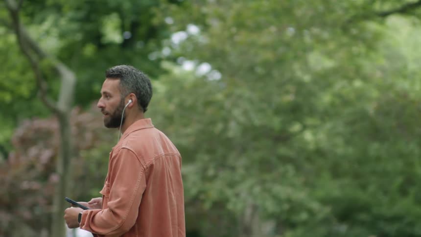 A man walks and talks outside while listening to an ear bud plugged into a smartphone
