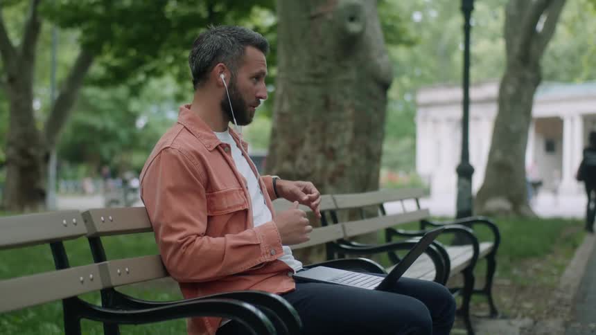 A man sits on a bench outdoors in a park having a video call on a laptop