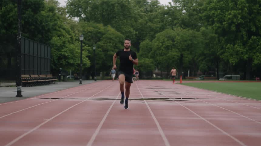 An athletic man runs on a red track surrounded by green trees in an urban park