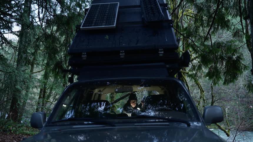 Man with a flashlight going through the trunk of a car that is parked next to a river in the forest