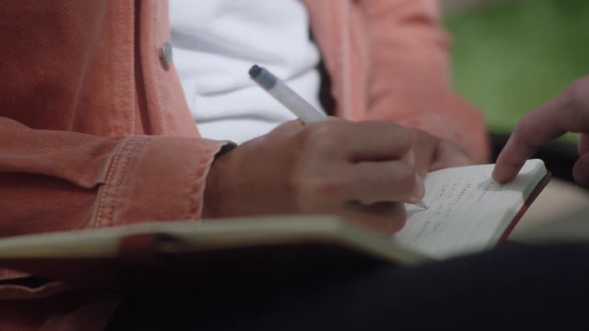 A person writes in a notebook outside while sitting on a park bench