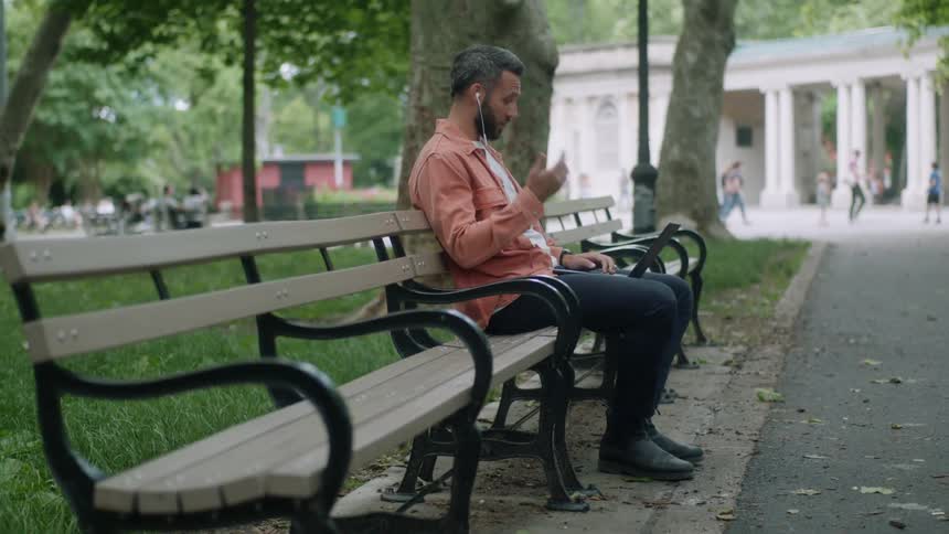 Man uses headphones during a conversation as he sits on a bench in a busy park and types on a laptop